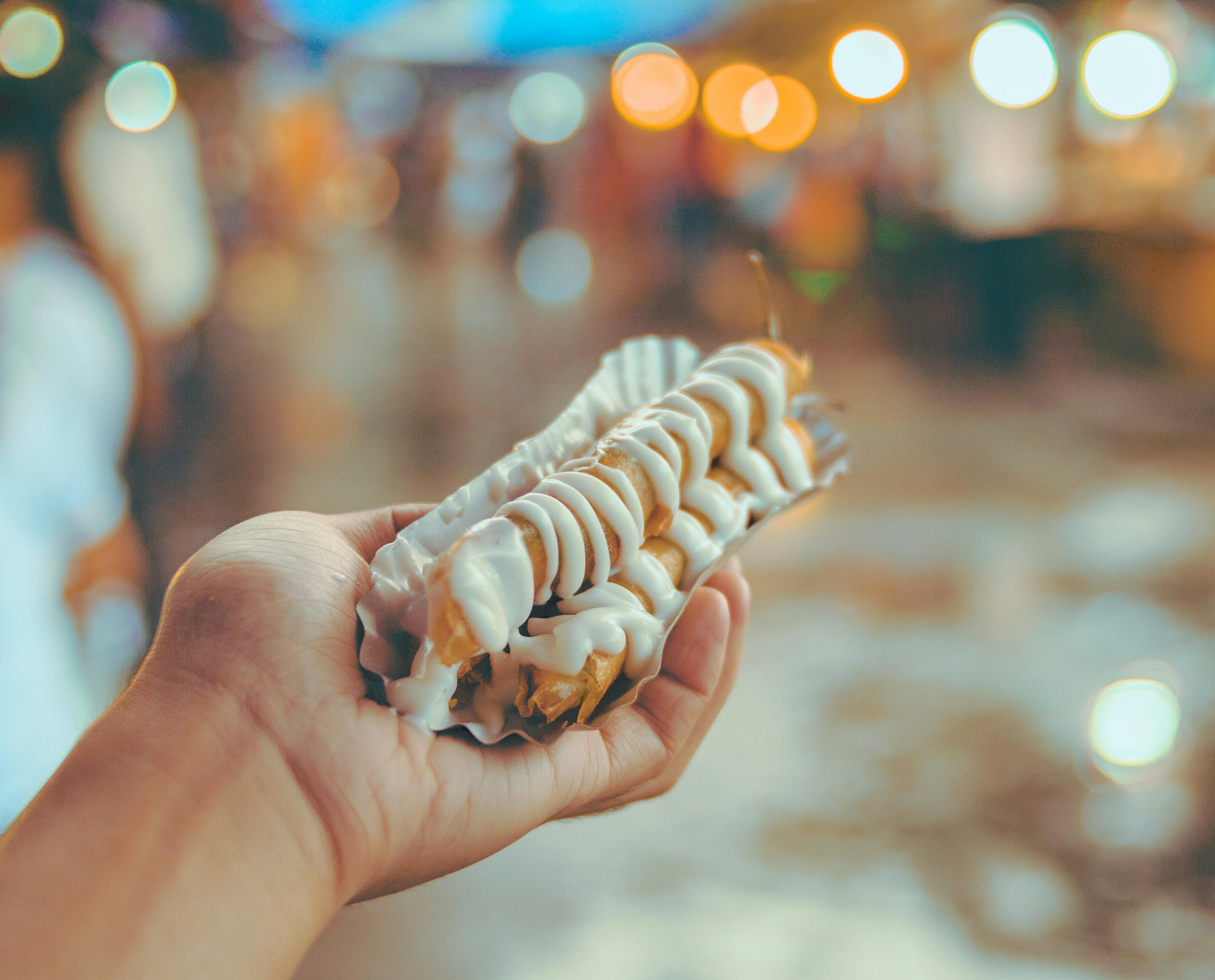 Street food in primo piano con abbondante maionese affumicata, tenuto in mano su sfondo sfocato e luminoso