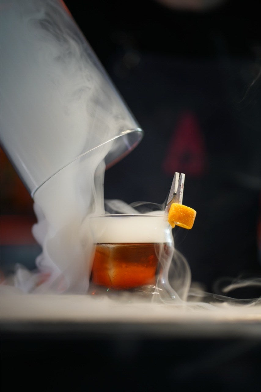 An amber cocktail surrounded by smoke beneath a glass dome